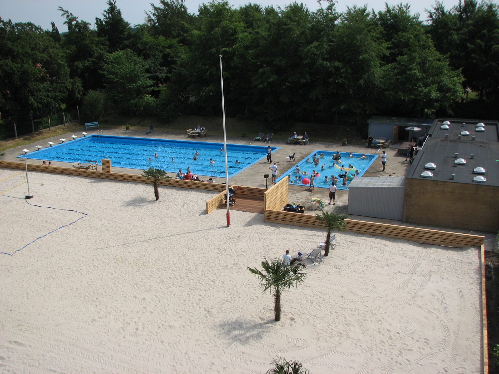 Overbliksbillede af et stort svømmebassin, et lille børnebassin og et stort sandareal med palmer og plads til leg og hvile. Samt en opmærket bane til beachvolley.